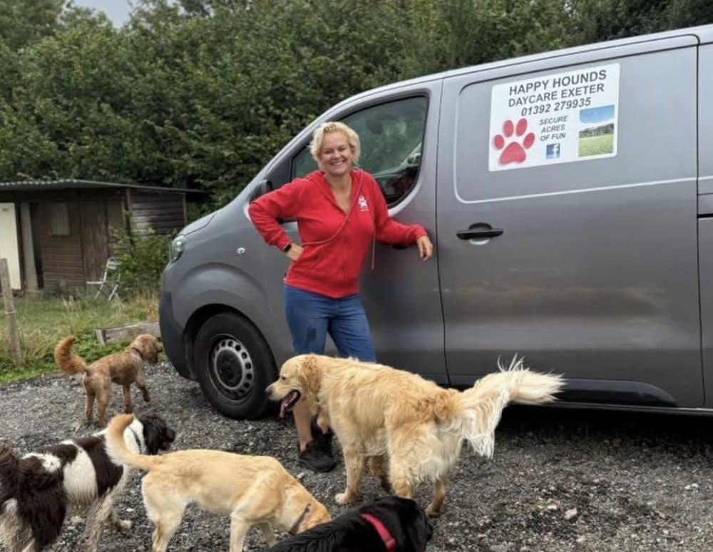 Hayley standing by the grey Happy Hounds van with the pack gathered round — six daycare dogs greeting her at the start of the day